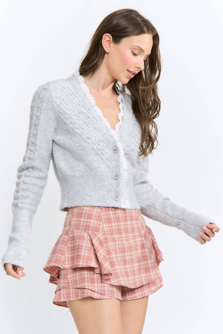 A woman modeling a light gray cable-knit cardigan with a scalloped V-neck and button-front closure, paired with a pink plaid ruffled mini skirt, against a plain white studio background.
