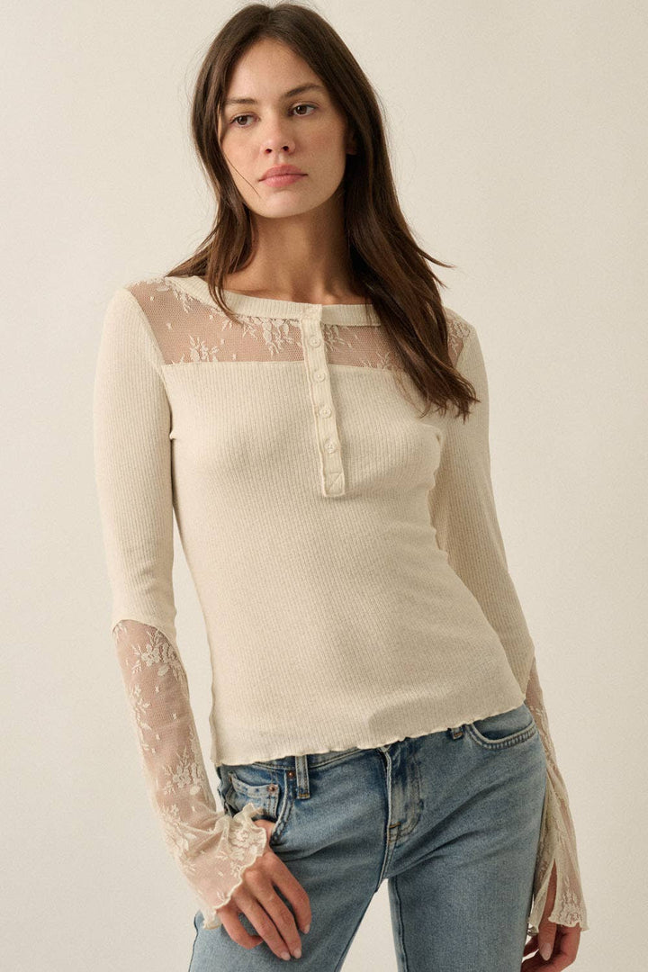 A woman modeling a cream ribbed knit long-sleeve top with sheer lace panels across the chest and on the sleeves, paired with blue jeans against a plain light beige background.