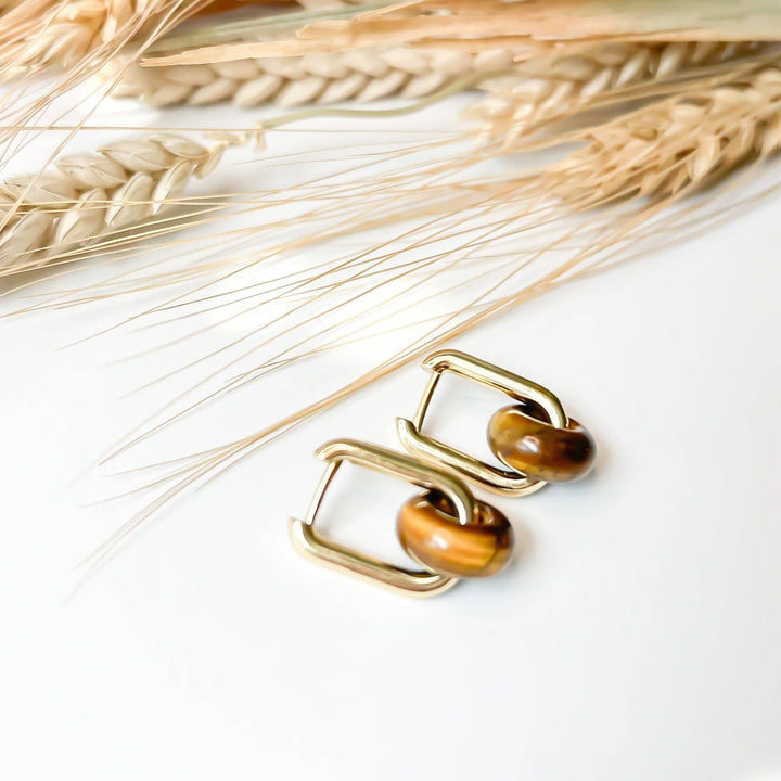 A pair of gold-toned geometric hoop earrings with brown tiger's eye beads, displayed on a white surface with dried wheat stalks in the background.