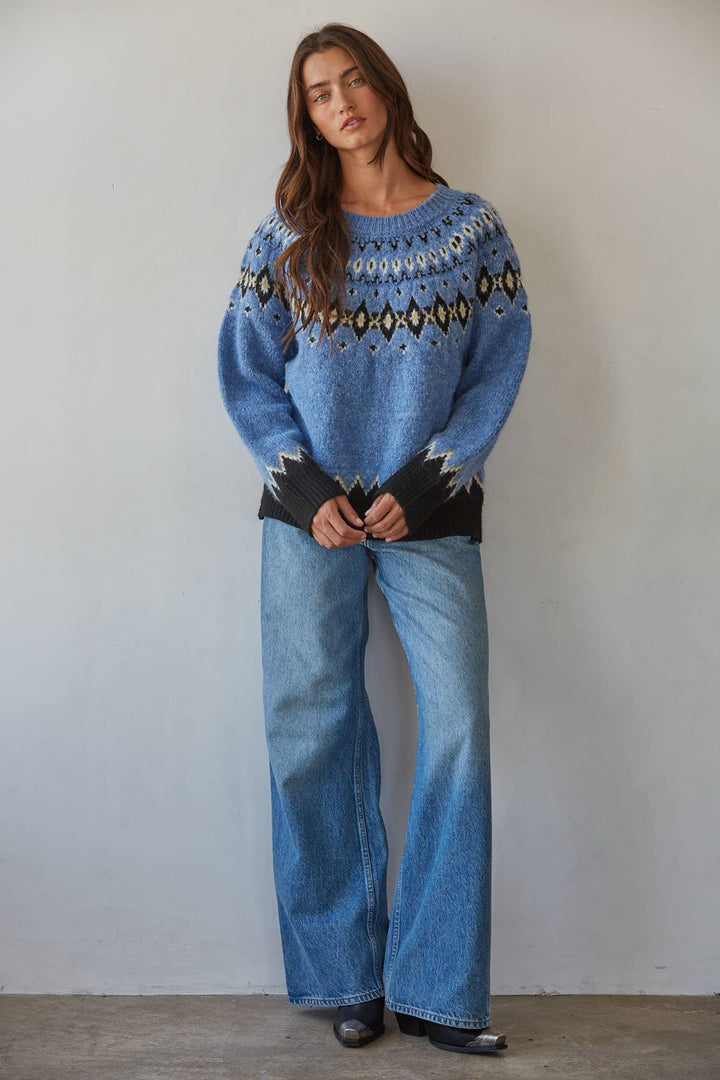 A woman stands against a plain light gray wall wearing a blue knit sweater with a geometric Fair Isle pattern across the chest and black cuffs, paired with wide-leg blue jeans and black shoes.