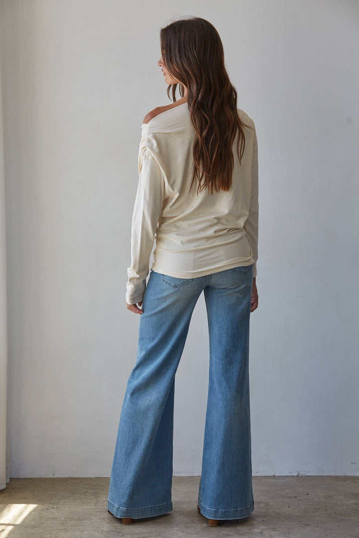 A woman is shown from behind wearing a cream off-the-shoulder basic long-sleeve top and light blue wide-leg jeans, standing against a plain light gray wall.