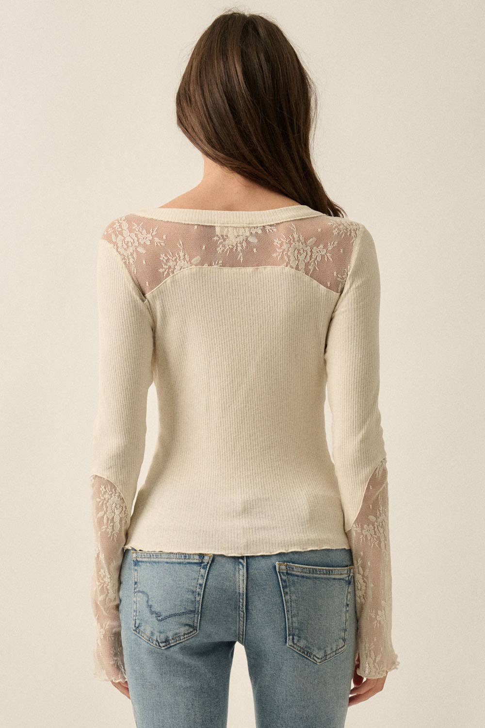 A woman viewed from the back wearing a cream ribbed knit henley top with sheer floral lace panels across the shoulders and down the sleeves, paired with light blue jeans against a plain off-white background.