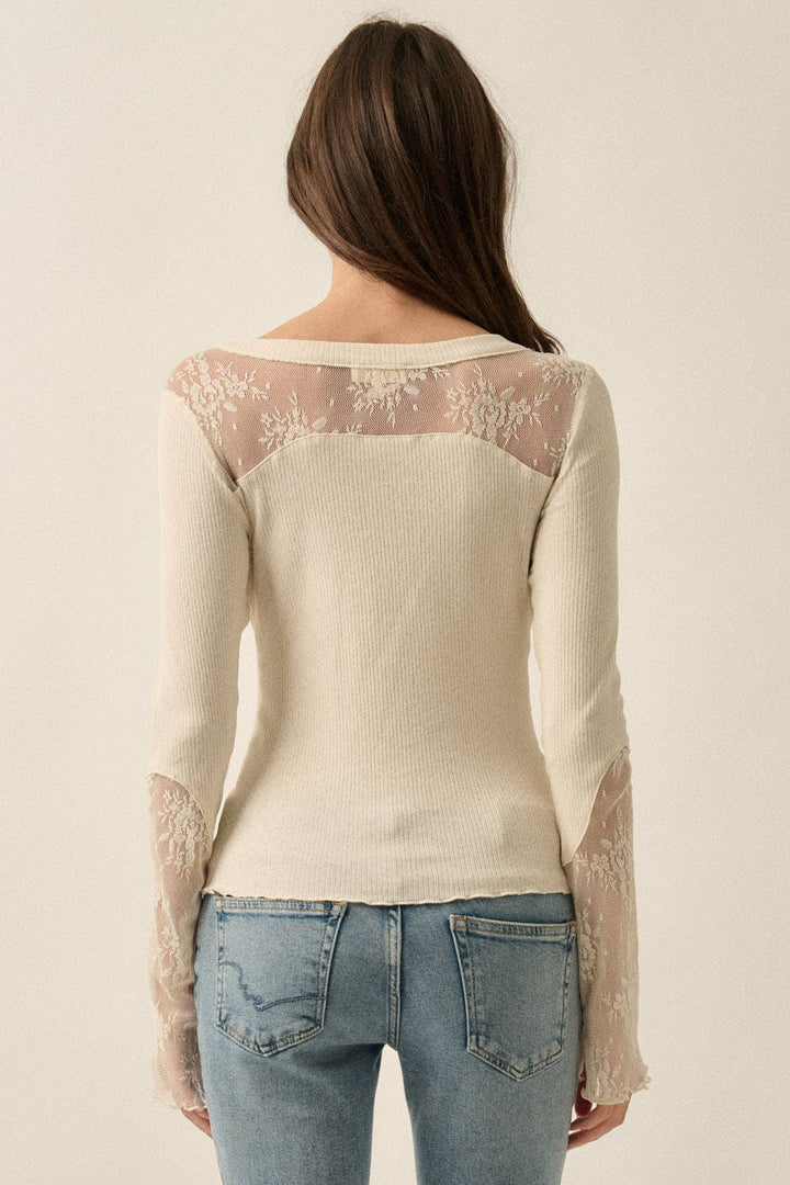 A woman viewed from the back wearing a cream ribbed knit henley top with sheer floral lace panels across the shoulders and down the sleeves, paired with light blue jeans against a plain off-white background.