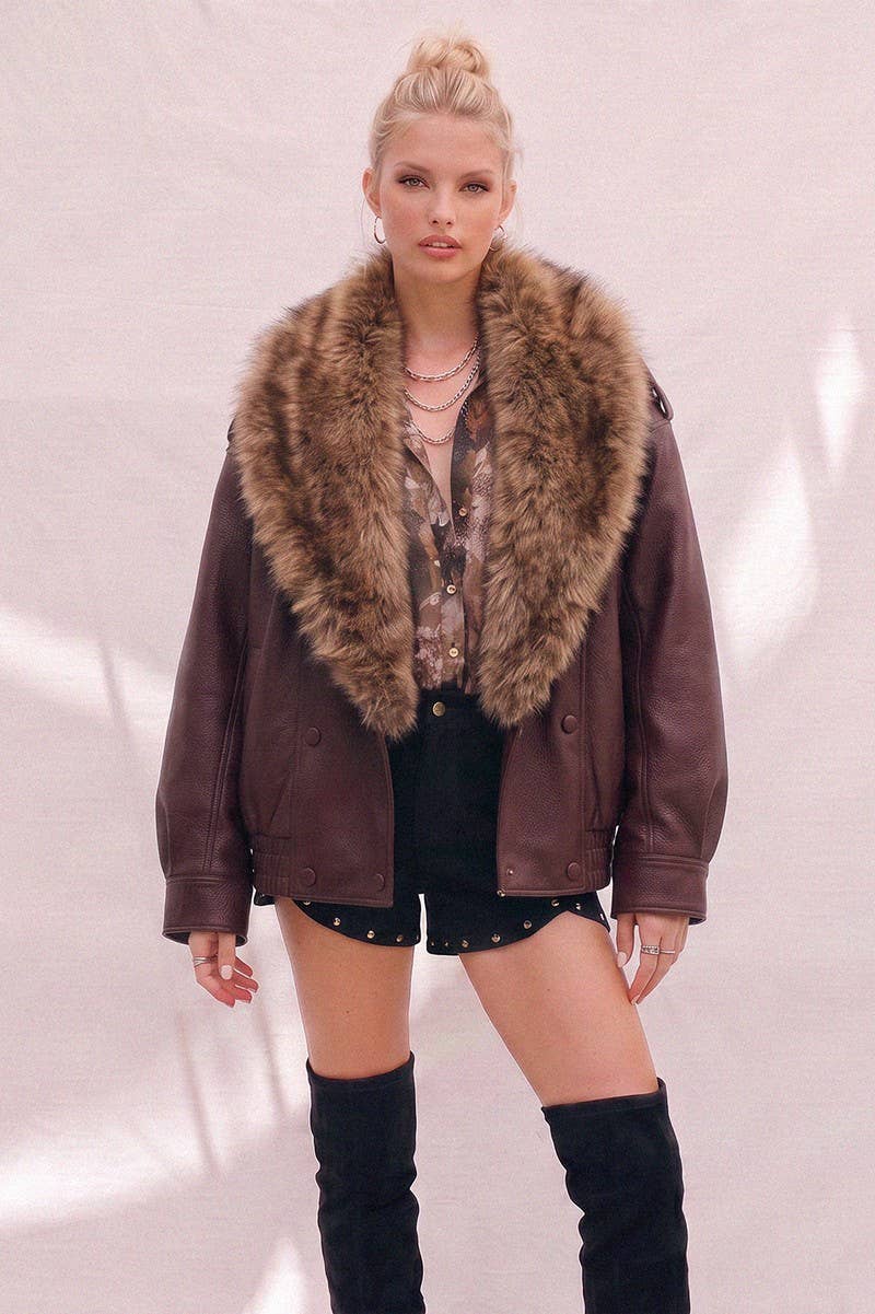 A model wearing a burgundy faux-leather aviator jacket with a large brown fur collar, styled with a patterned top, black studded shorts, and black over-the-knee boots against a light pink textured wall.