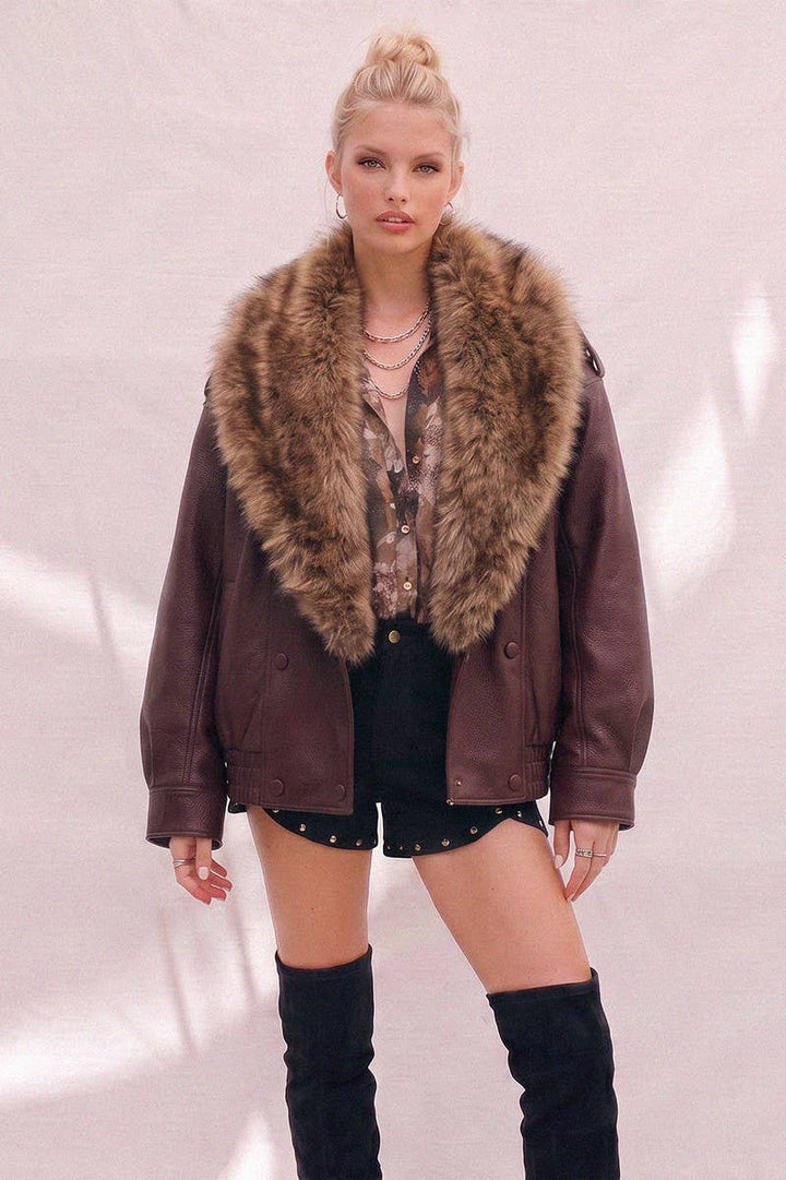 A model wearing a burgundy faux-leather aviator jacket with a large brown fur collar, styled with a patterned top, black studded shorts, and black over-the-knee boots against a light pink textured wall.