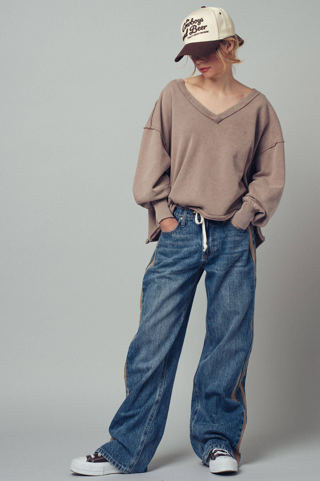 Person wearing a beige sweatshirt, blue jeans, and a cap on a gray background