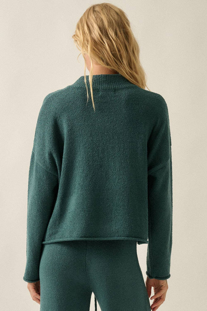 A woman shown from the back wearing a dark teal green knit sweater and matching knit pants against a plain light beige studio background.