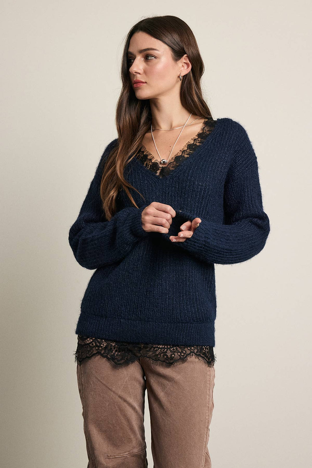 A woman wearing a dark navy blue ribbed knit sweater with a lace V-neckline, paired with brown pants, standing against a plain beige studio background.