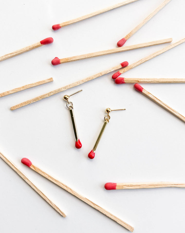 Gold matchstick earrings with red tips on a white background with scattered matchsticks.