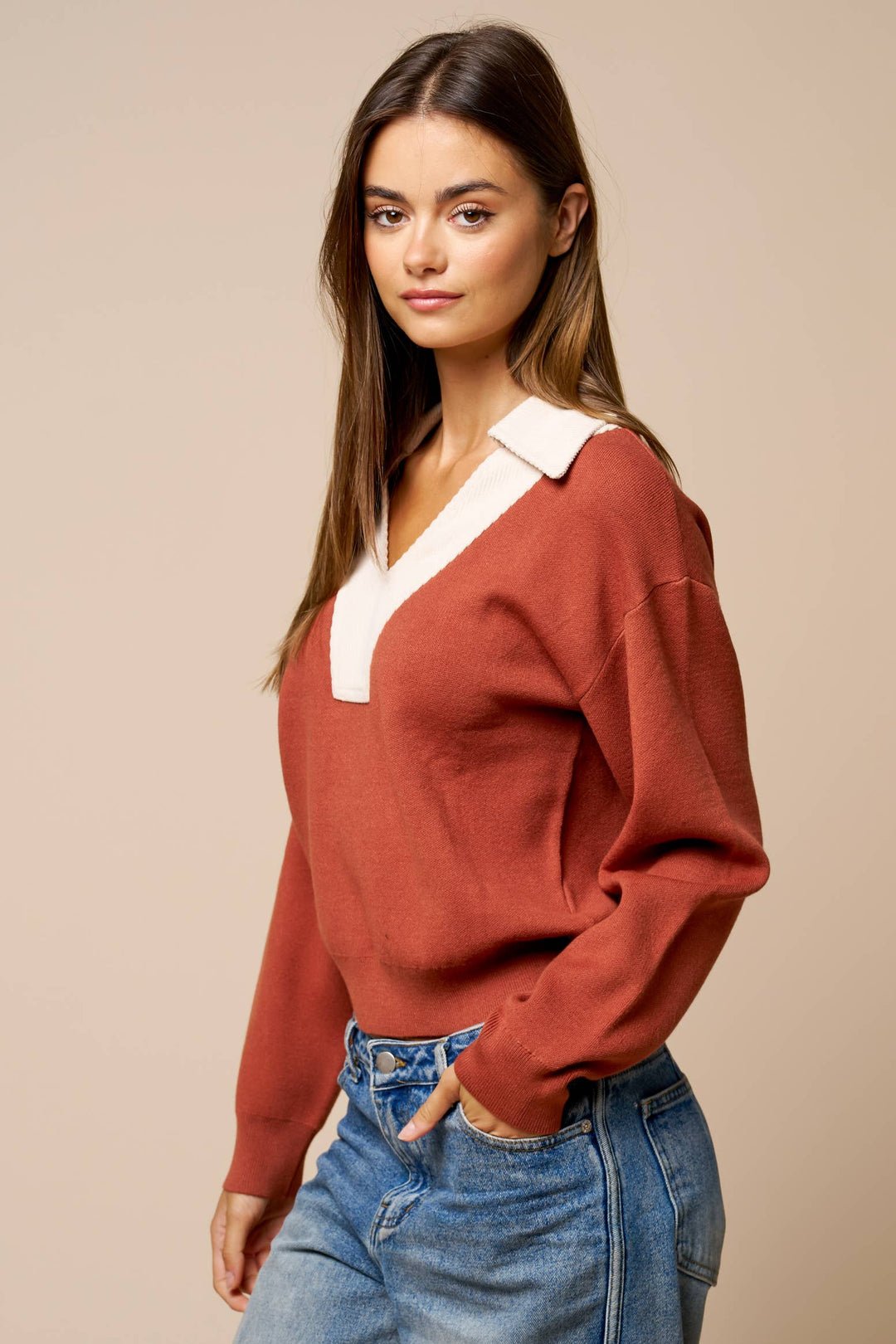 A woman modeling a rust-colored knit sweater with a contrasting white V-neck collar, paired with blue jeans, against a solid beige studio background.