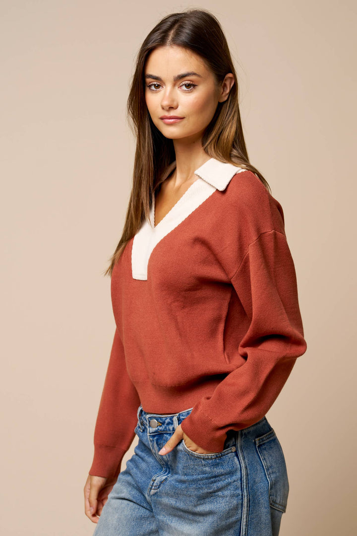 A woman modeling a rust-colored knit sweater with a contrasting white V-neck collar, paired with blue jeans, against a solid beige studio background.