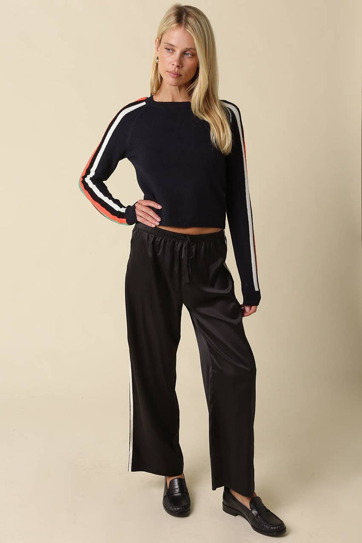 A woman modeling a navy long-sleeve crop top with white and orange stripe details on the sleeves, paired with black wide-leg pants and black loafers, against a solid beige background.
