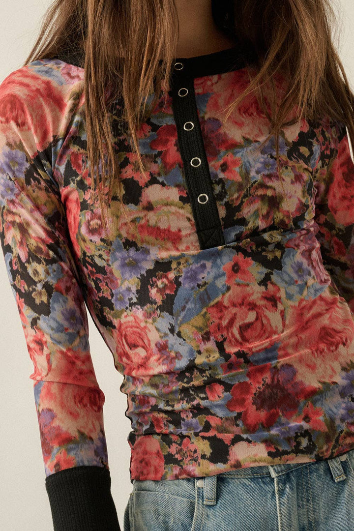 A close-up of a woman wearing a long-sleeve floral top with a black collar and three silver grommets, paired with blue jeans.