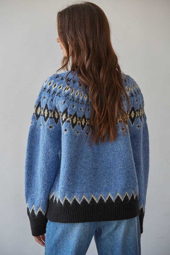 A woman viewed from the back wearing a fuzzy light blue knit sweater with a dark navy and beige geometric pattern across the shoulders and a matching dark navy hem, paired with blue jeans against a plain light gray wall.
