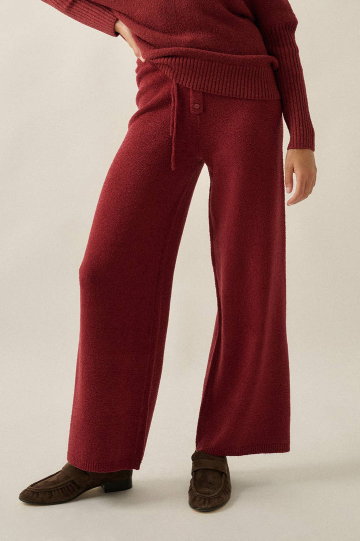 A person wearing a ribbed knit sweater and matching wide-leg trousers in a deep burgundy red, paired with dark brown loafers, against a plain light beige studio background.