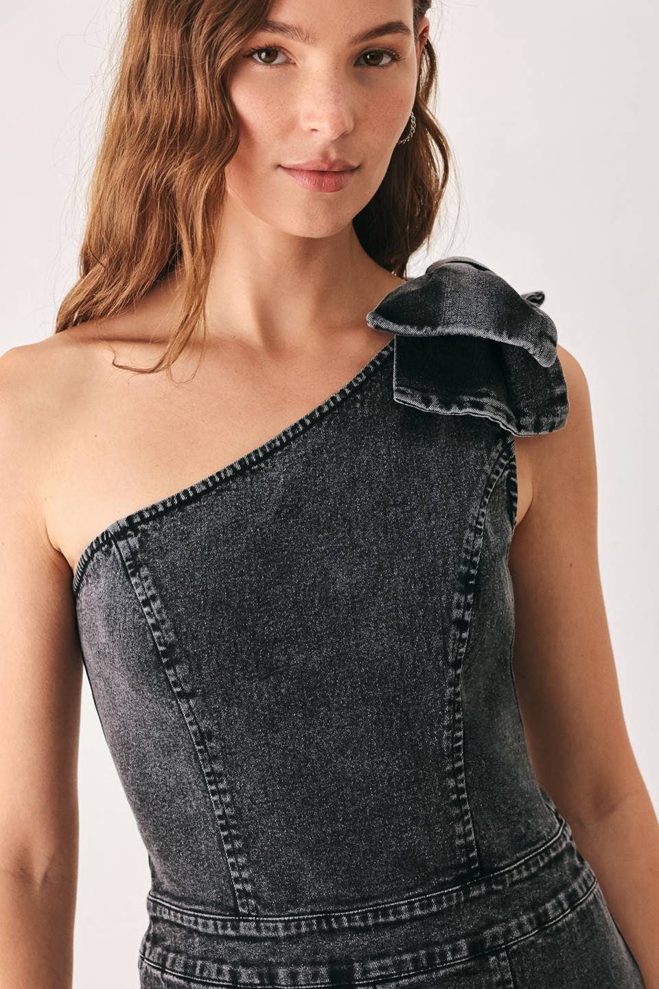 A close-up of a model wearing a dark gray washed denim one-shoulder top with a ruffled bow detail on the shoulder, set against a plain light background.