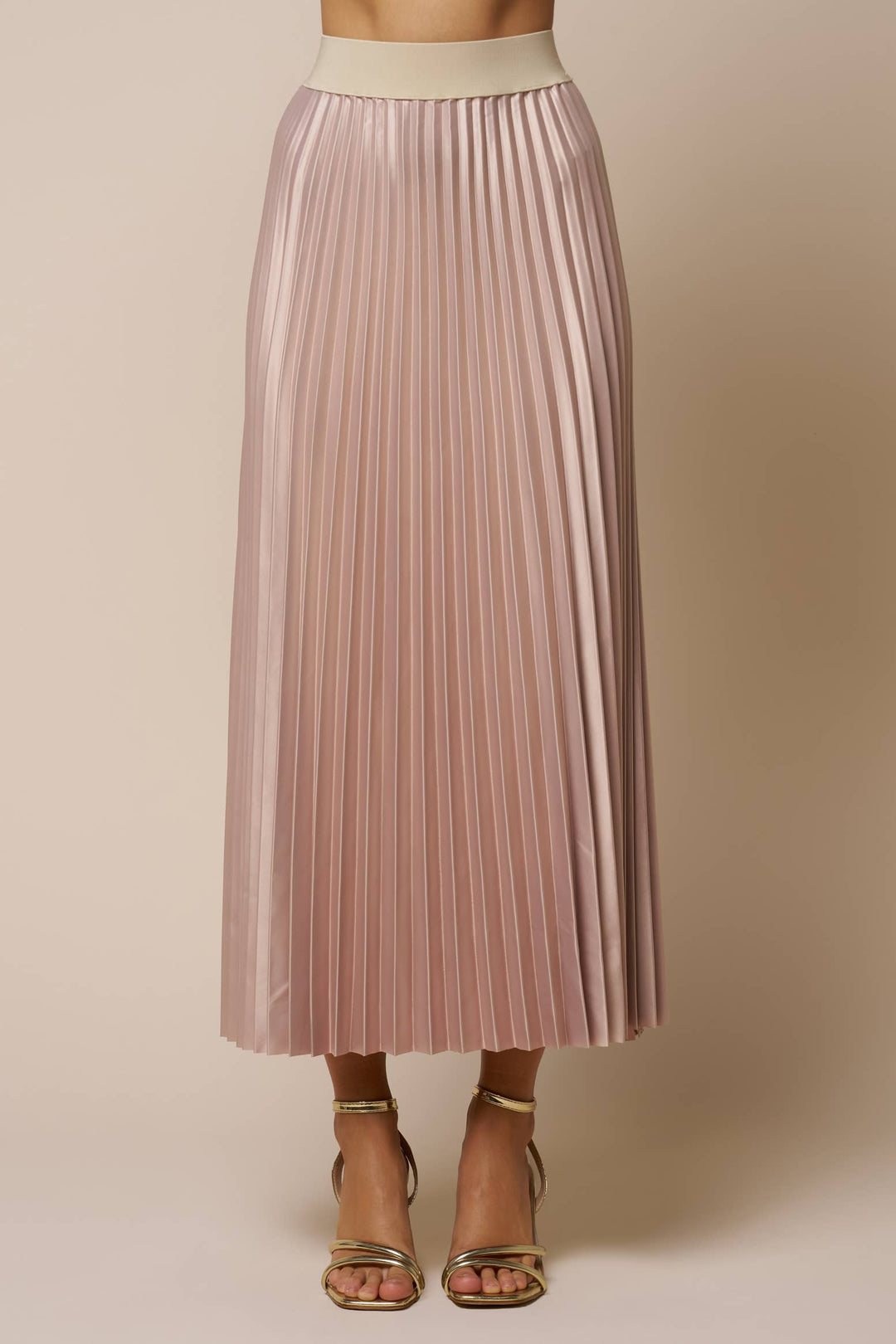 A pleated midi skirt in a champagne color, shown on a model from the waist down wearing gold heeled sandals against a solid beige background.