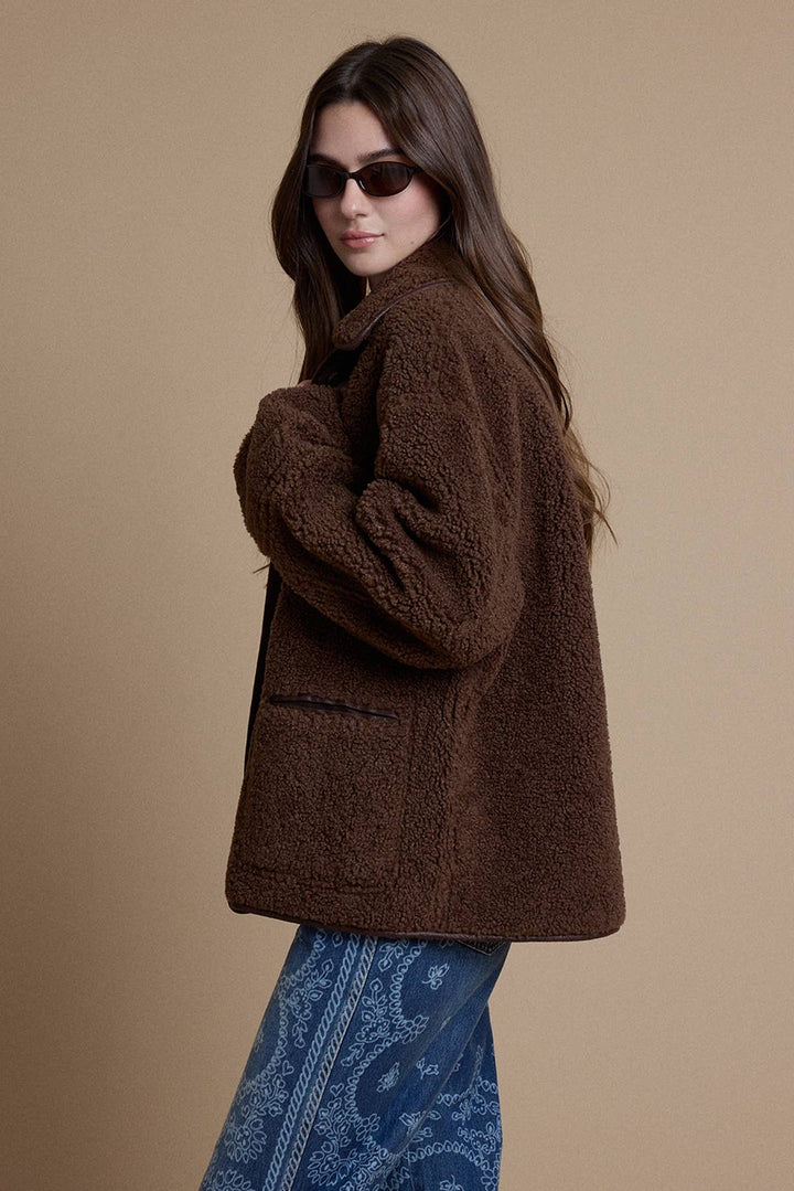 A woman wearing a brown sherpa fleece jacket and blue paisley pants, photographed against a solid beige studio background.