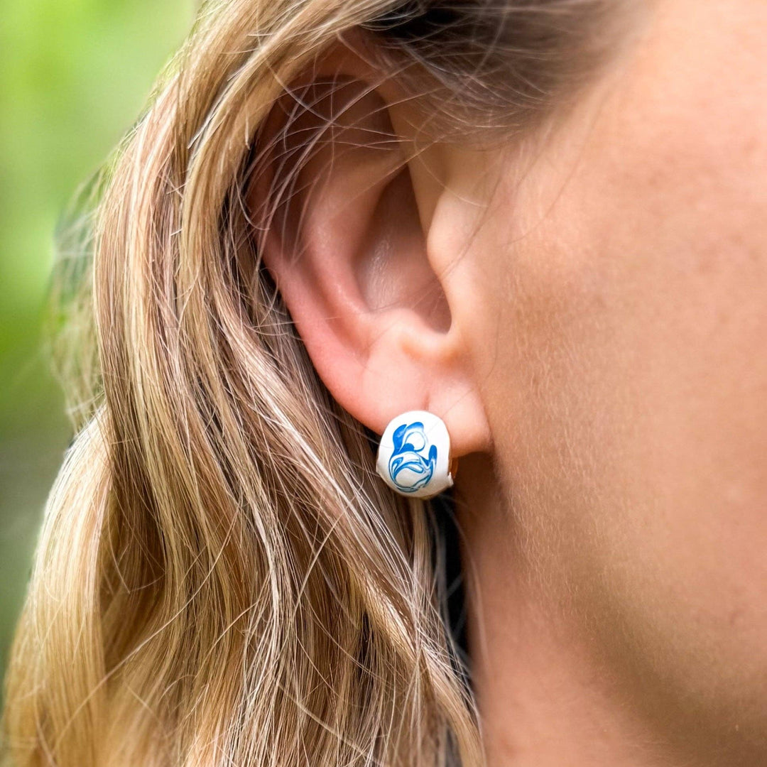 Close-up of an ear wearing a small white and blue hoop earring with a blurred natural background