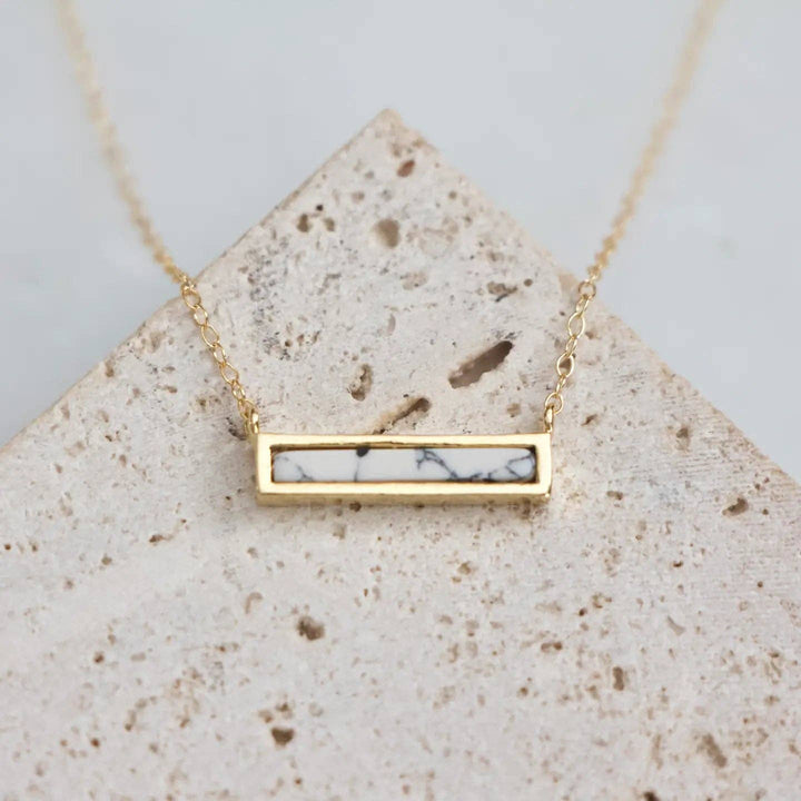 Gold necklace with a marbled dainty bar pendant on a textured stone surface