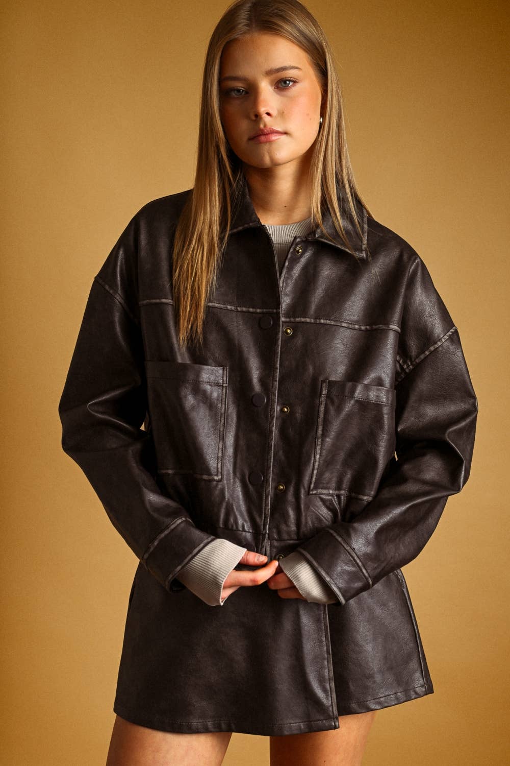 A woman wearing an oversized dark brown faux-leather button-up shirt jacket with contrast stitching, photographed against a solid warm tan studio background.