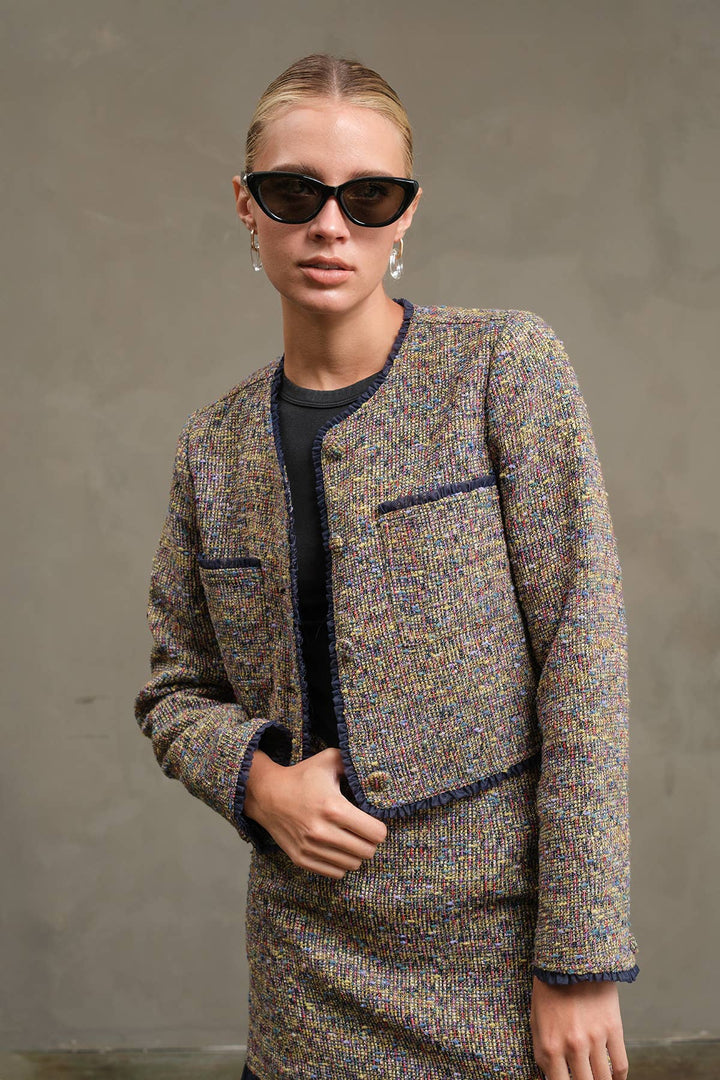 A model wearing a tweed jacket and matching skirt in a brown, beige, and blue patterned weave, styled with a black top, black sunglasses, and silver hoop earrings against a textured gray wall.