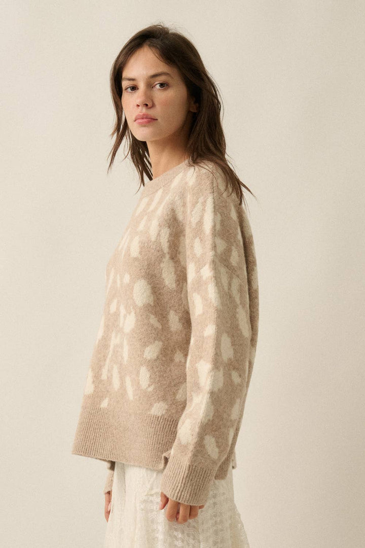 A woman wearing a beige knit sweater with a white abstract spotted pattern, photographed against a plain light beige studio background.