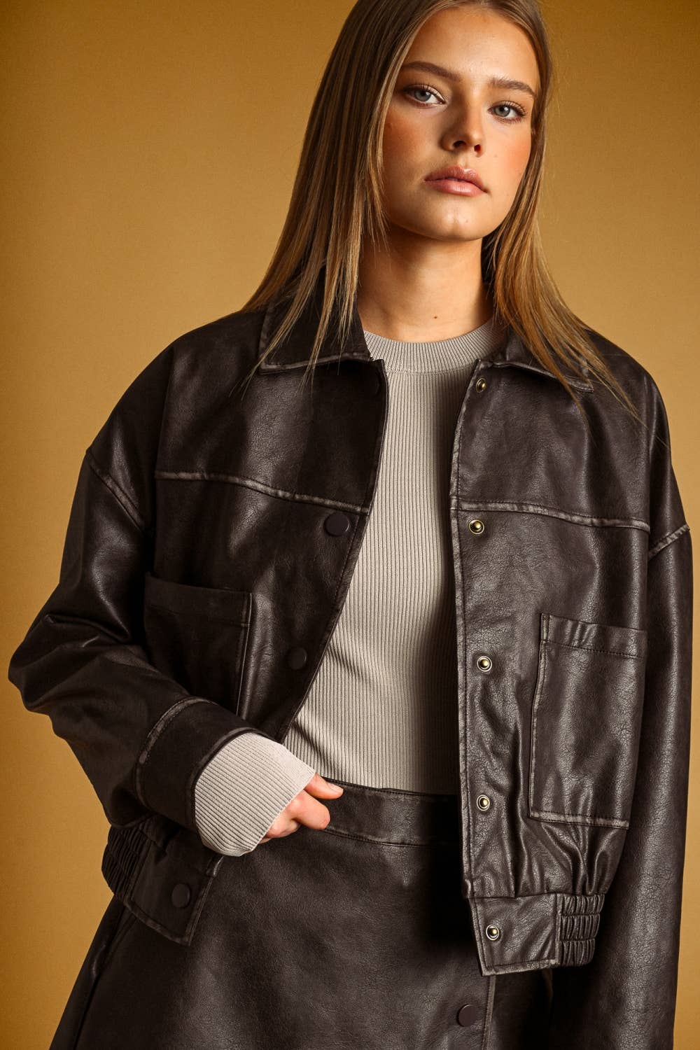 A woman wearing a dark brown faux-leather utility jacket over a ribbed gray top, posed against a solid warm brown background.