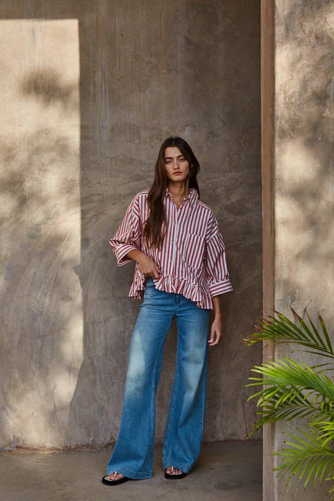 Woman wearing a red and white striped short sleeve shirt and blue jeans standing against a textured wall with a plant in the corner.