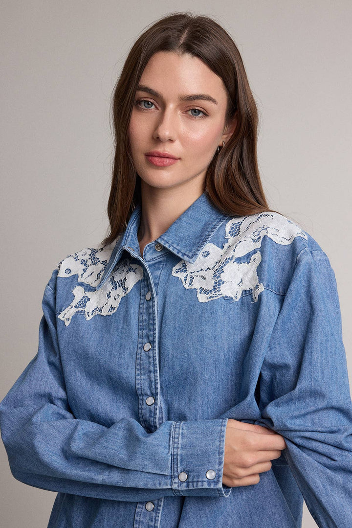 A woman wearing a blue denim button-up shirt with white floral lace embroidery across the shoulders, posed against a neutral light gray background.