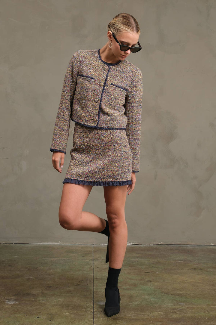 A model wearing a tweed mini dress set consisting of a cropped jacket and a matching mini skirt, in a beige and brown pattern with navy blue trim, styled with black ankle boots and sunglasses against a textured gray studio backdrop.