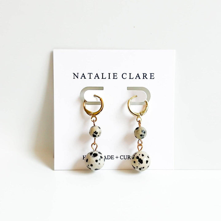 A pair of Natalie Clare dangle earrings displayed on a white branded card. The earrings feature gold-tone hoop tops, small round beads, and larger speckled white beads with black spots, resembling a dalmatian pattern.
