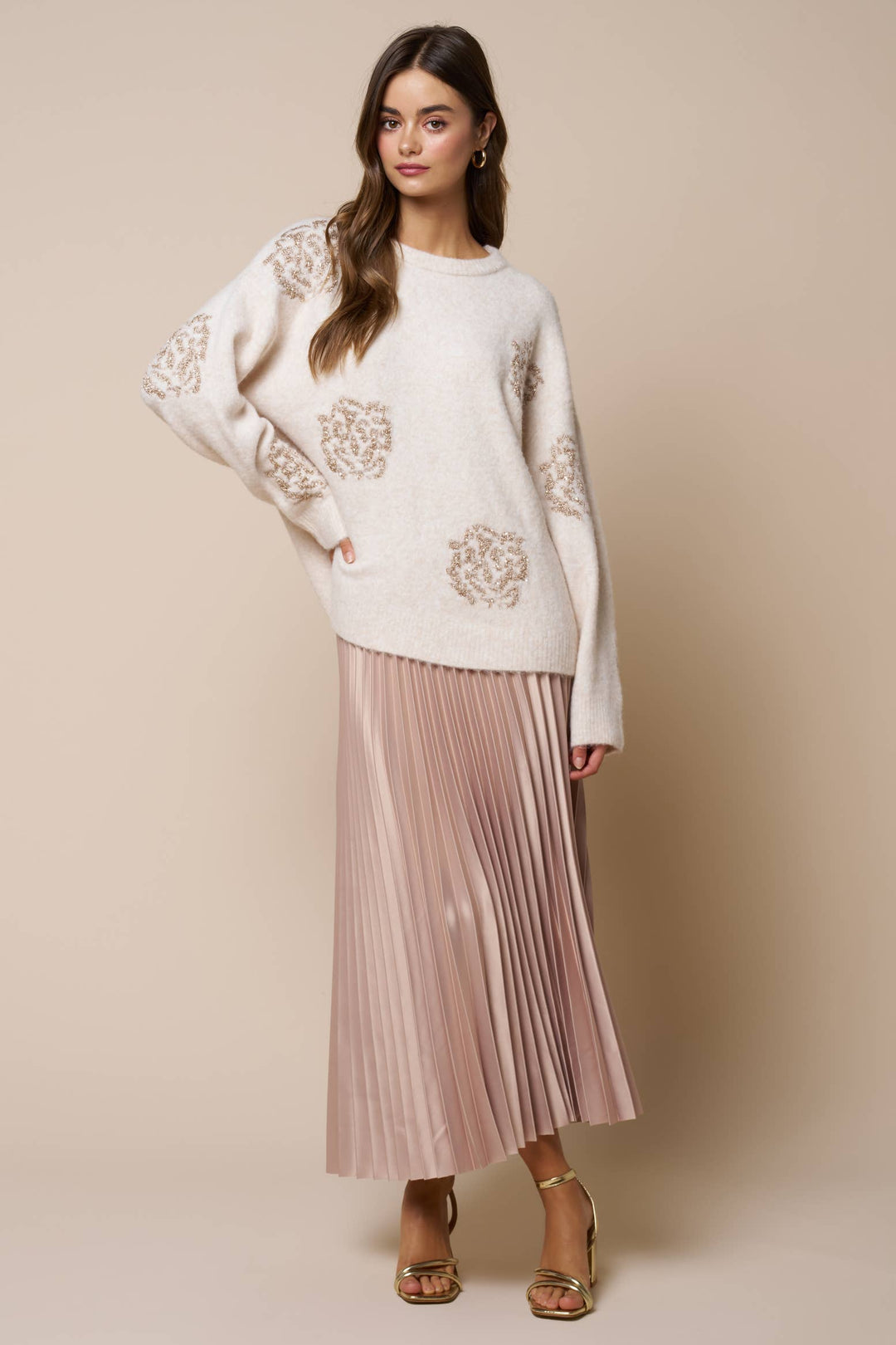 A woman modeling a cream knit sweater with beige floral embroidery and a dusty pink pleated midi skirt, paired with brown strappy heels, against a solid beige studio background.