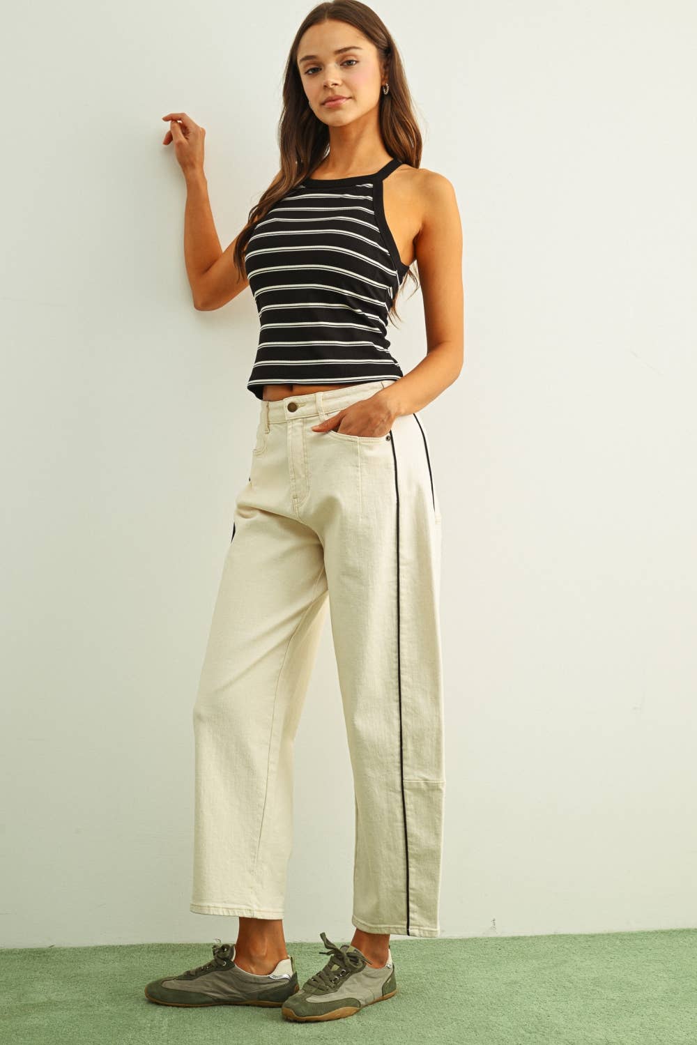 Woman wearing a striped tank top and beige pants against a white wall.