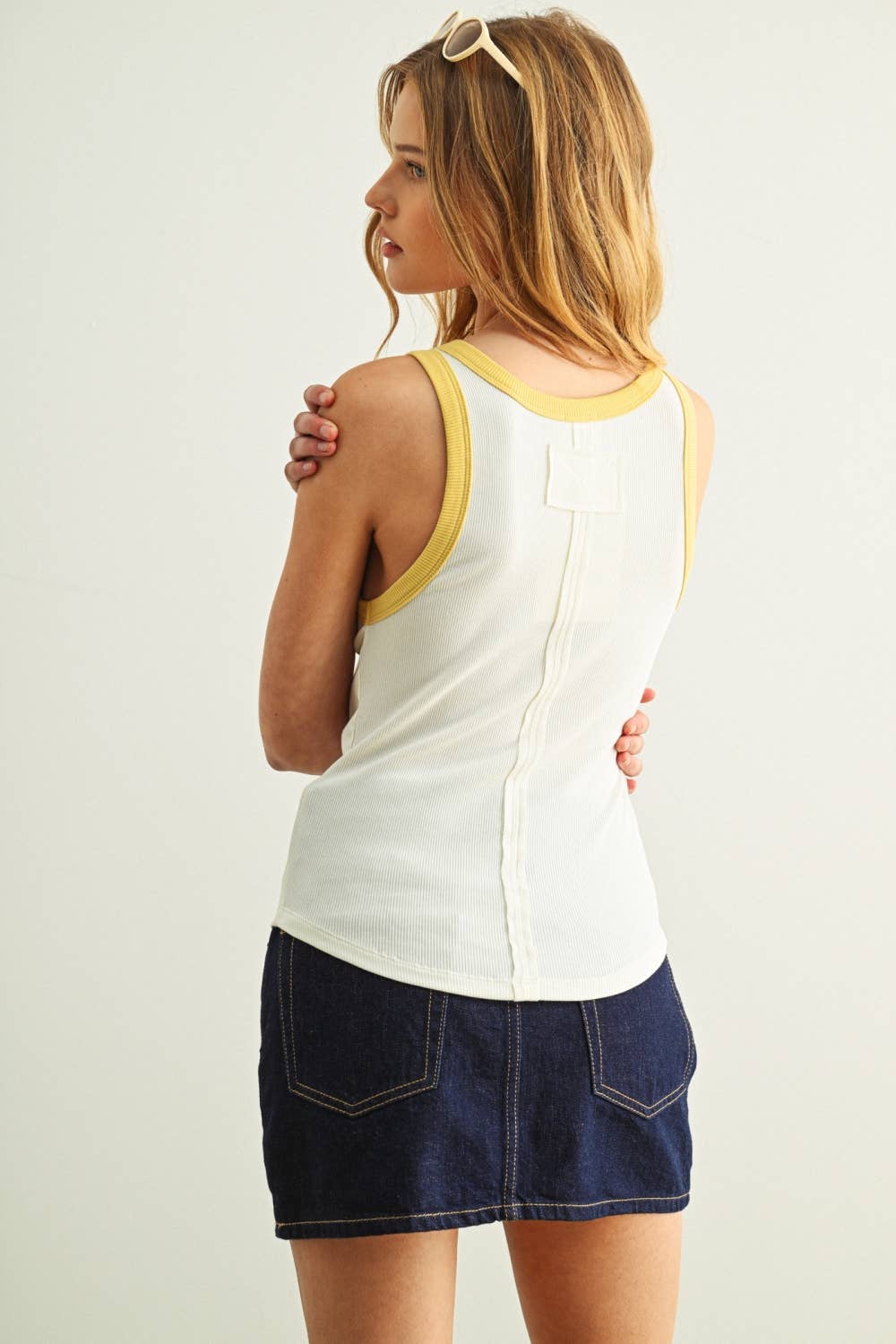 Woman wearing a white tank top with yellow trim and a denim skirt on a plain background