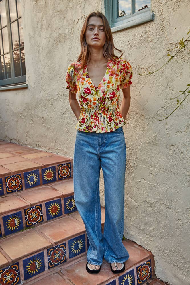 Woman wearing a colorful floral yellow and red short sleeve blouse and blue jeans standing on decorative steps.