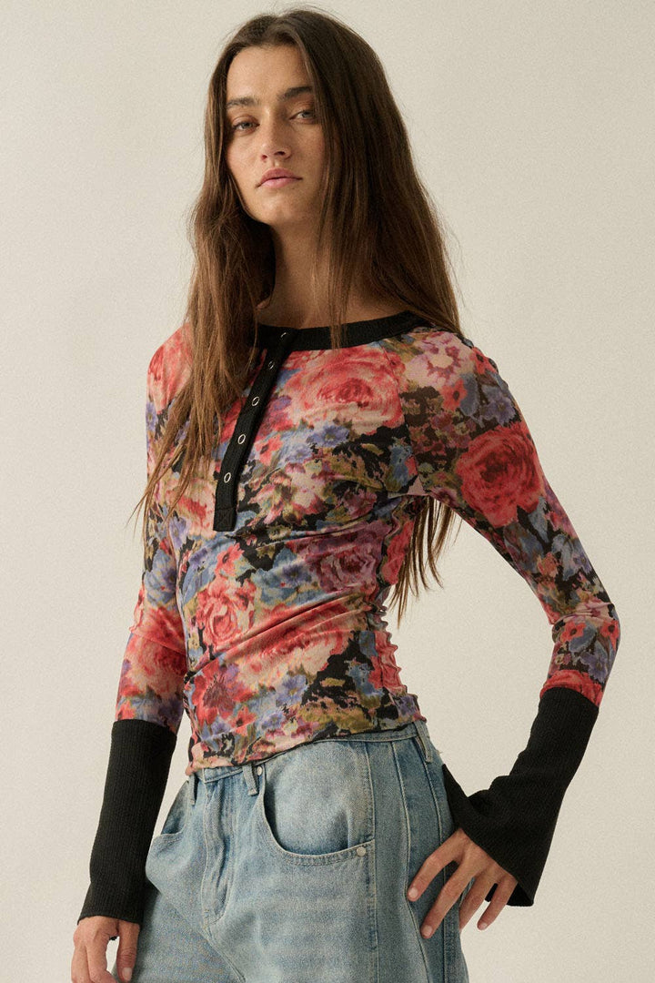 A woman modeling a long-sleeve floral top with black trim and black ribbed cuffs, paired with light blue jeans, against a plain light beige background.