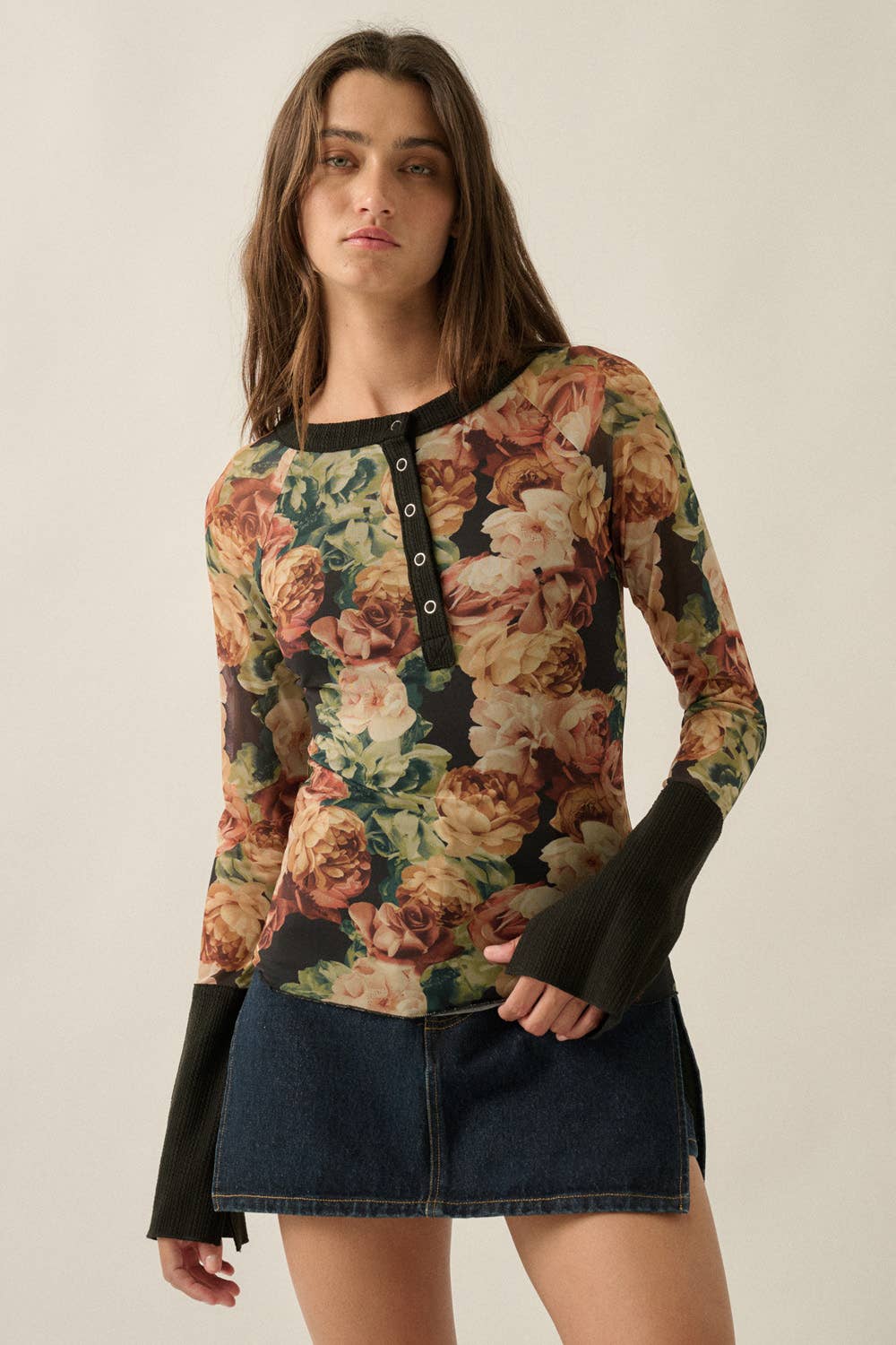 A woman modeling a long-sleeve floral knit top with black trim and button placket, paired with a dark denim skirt, against a plain beige studio background.