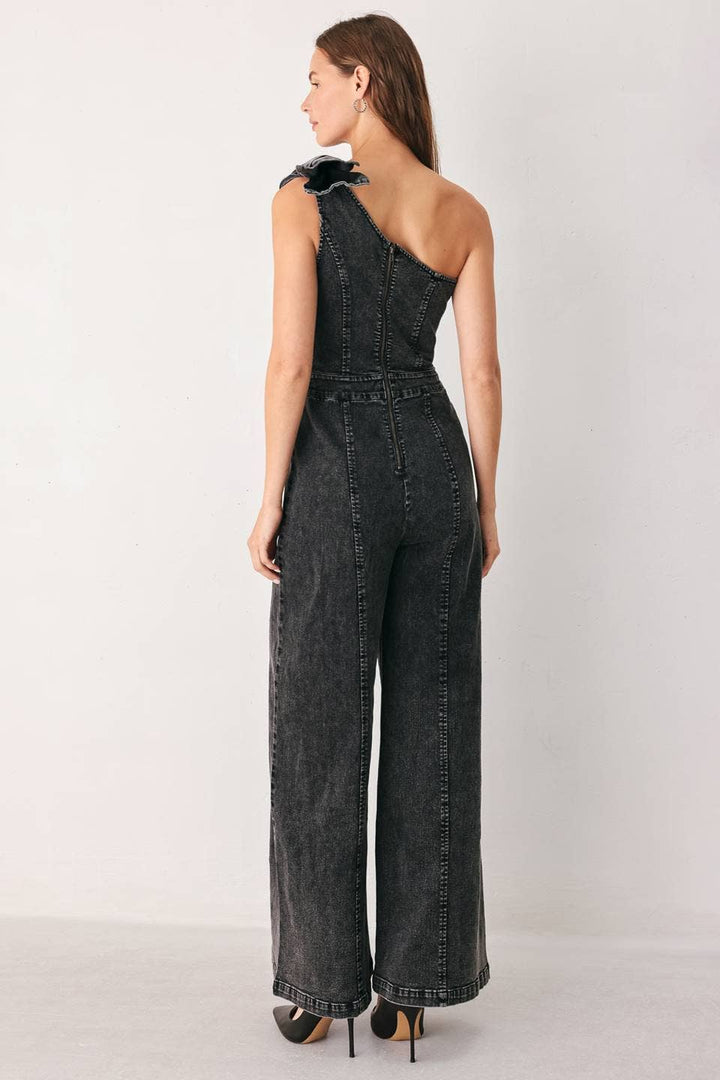 A woman wearing a dark gray denim one-shoulder jumpsuit with a bow shoulder detail, shown from the back against a plain light gray studio background.