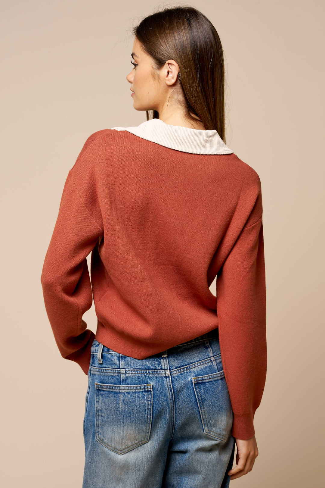 A woman shown from the back wearing a rust-colored knit sweater with a contrasting white collar, paired with blue jeans, against a solid beige background.