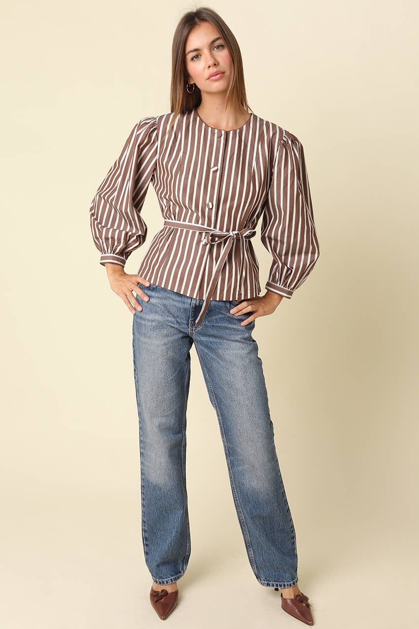 A woman modeling a brown and white vertically striped long-sleeve blouse with puff sleeves and a tie waist, paired with blue jeans and brown pointed shoes, against a solid beige background.