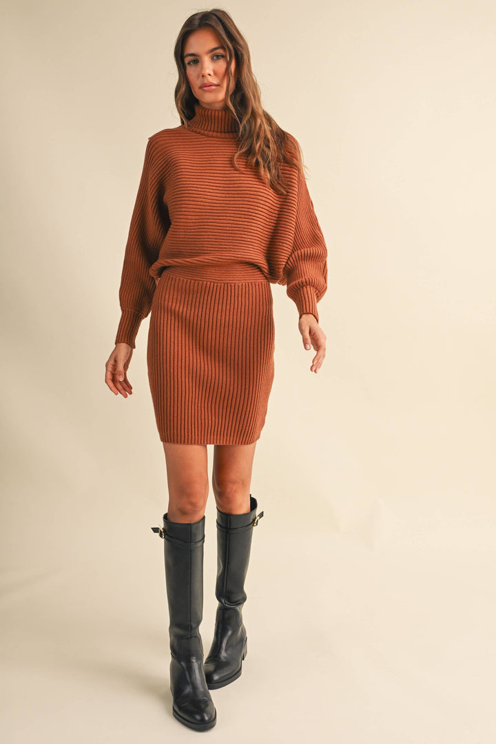 A woman modeling a rust-orange ribbed knit mock-neck sweater dress and paired with black knee-high boots, against a solid beige studio background.