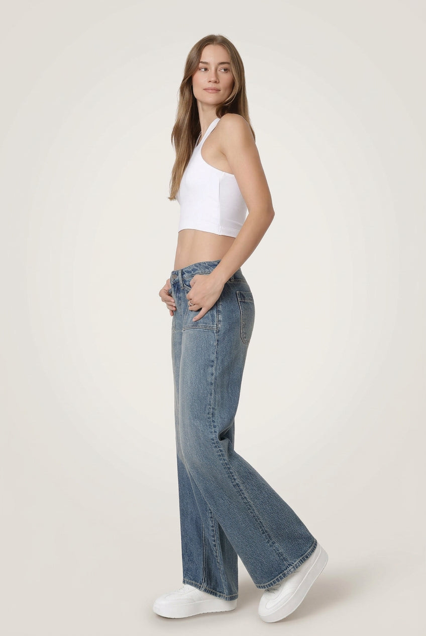 A woman modeling a pair of wide-leg blue jeans with a white crop top and white sneakers, standing against a plain white studio background.