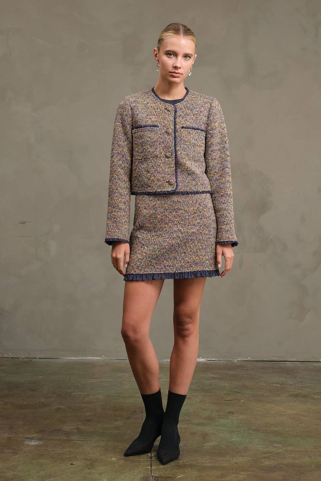 A model stands against a textured gray studio backdrop wearing a tweed mini dress with dark piping and fringe, paired with black ankle boots.