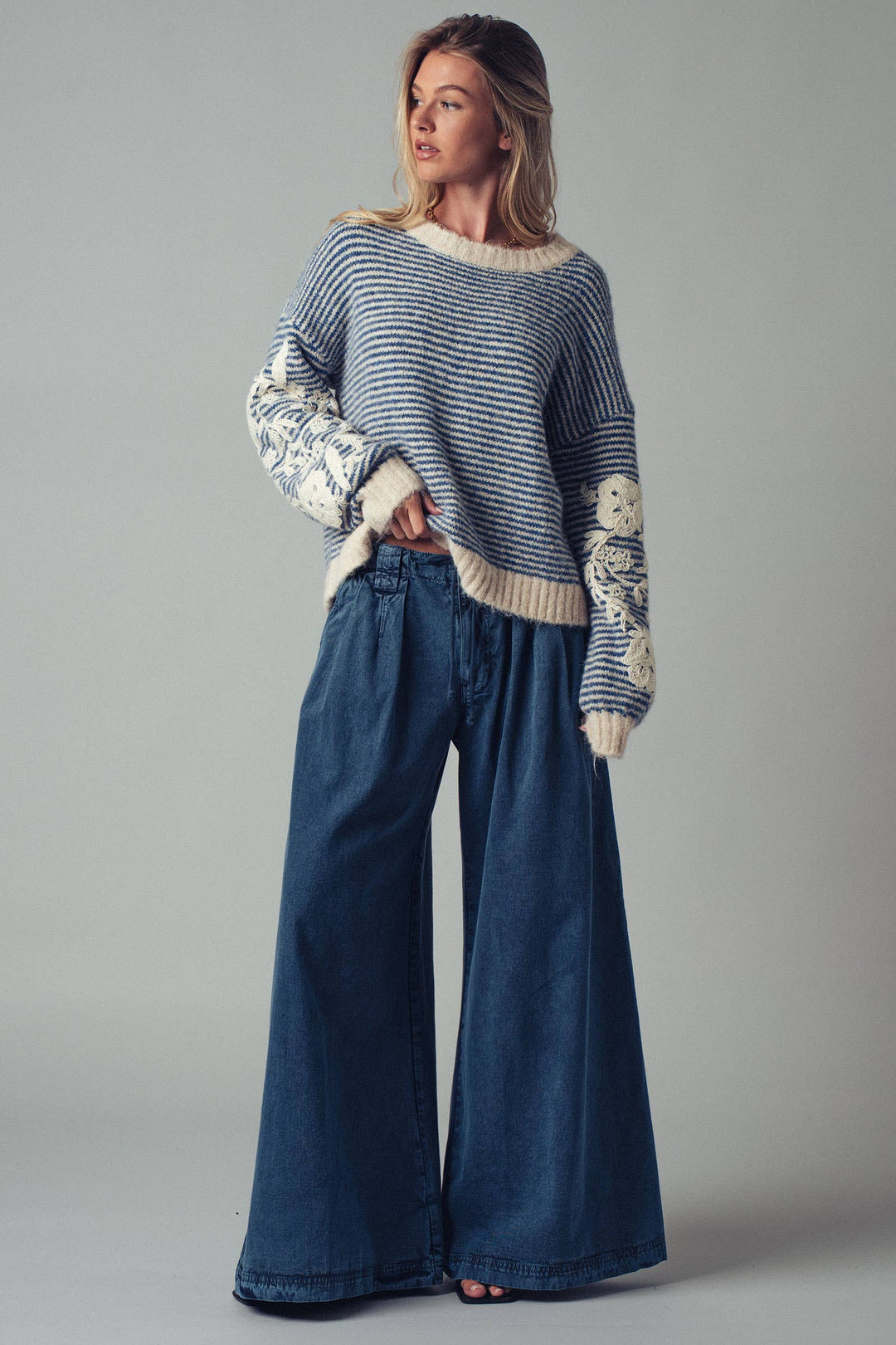 Woman wearing a striped sweater with floral crochet sleeve detail and wide-leg jeans on a gray background