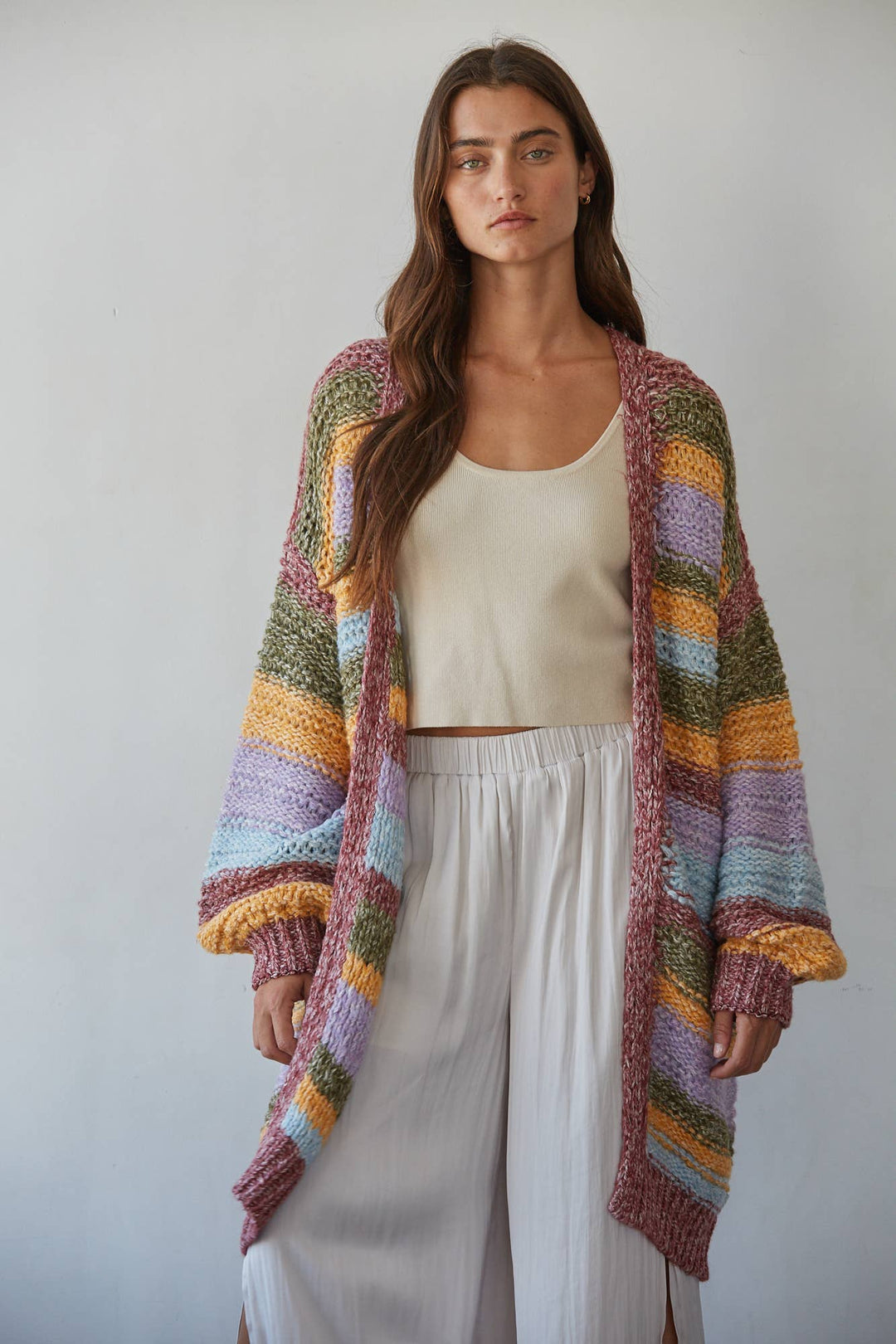 A woman modeling a chunky, open-front knit cardigan with horizontal stripes in pink, purple, blue, green, yellow, and orange, worn over a cream crop top and white wide-leg pants against a light gray background.