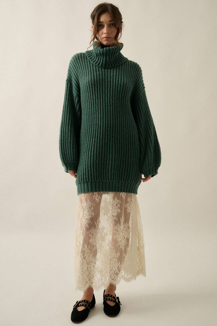 A model stands against a plain light beige studio background wearing an oversized dark green ribbed turtleneck sweater tunic, a cream lace midi skirt, and black flats with gold chain detailing.