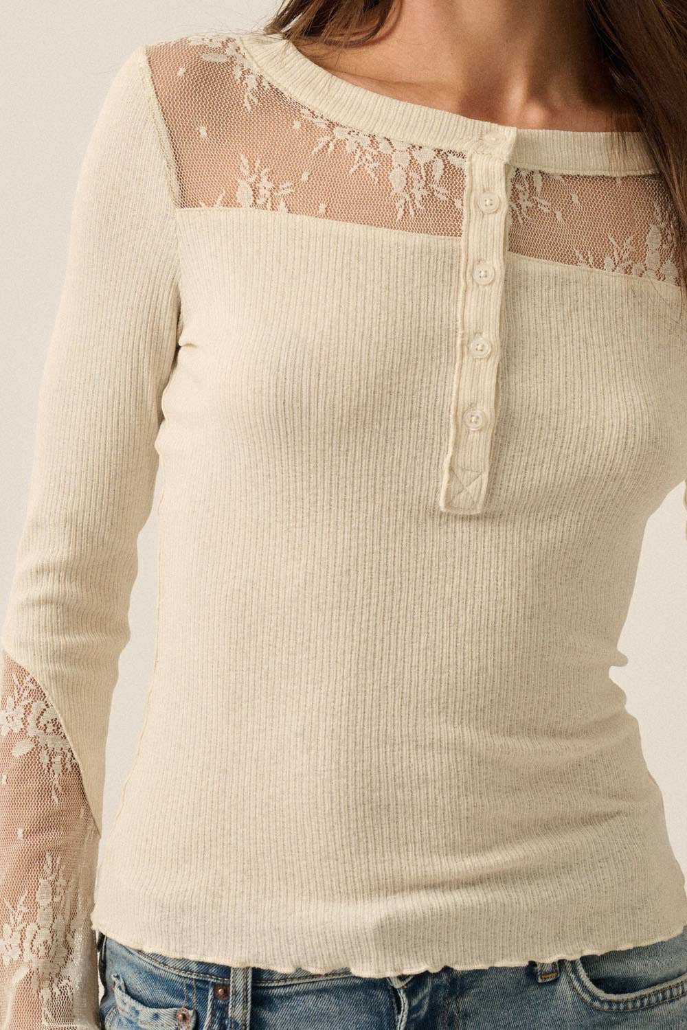 A close-up of a woman wearing a ribbed long-sleeve top in cream or off-white. The top features sheer floral lace shoulder panels and matching lace cuffs, with a short button placket at the neckline.