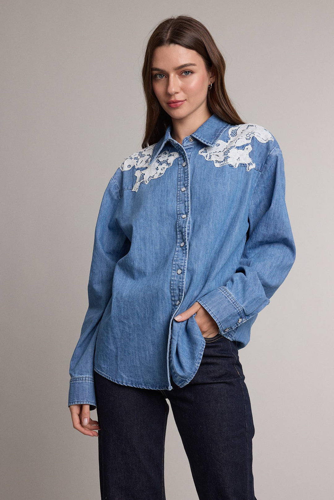 A woman modeling a blue denim shirt with white floral embroidery on the shoulders, paired with dark jeans, against a solid gray studio background.