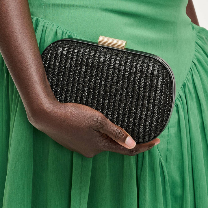 Person holding a black woven clutch purse against a green dress background